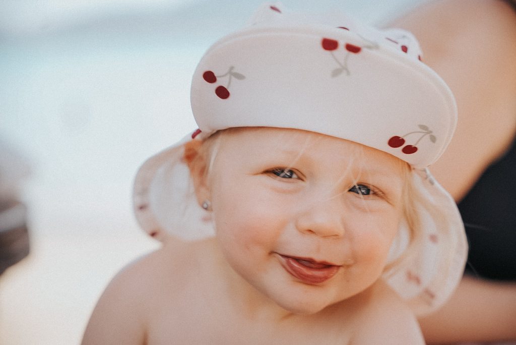 Lachendes Baby blickt mit einem hellen Kopftuch mit Kirschmuster direkt in die Kamera.