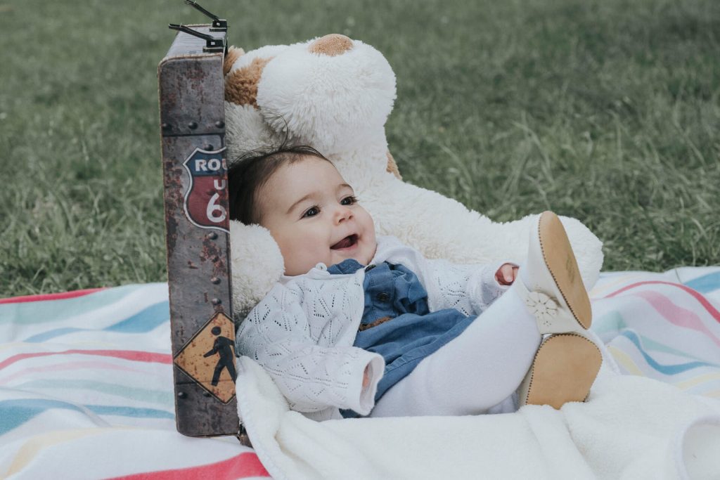Lachendes Baby liegt mit einem großen Stofftier auf einer Picknickdecke.