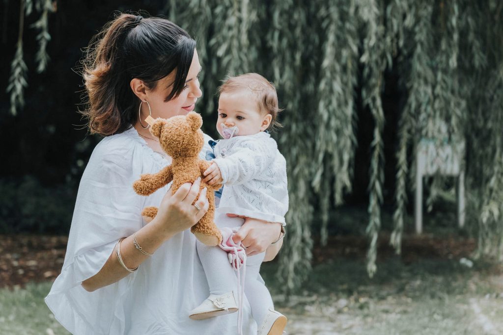 Mutter hält ihr Baby mit Stofftier im Arm vor einem grünen Baum.