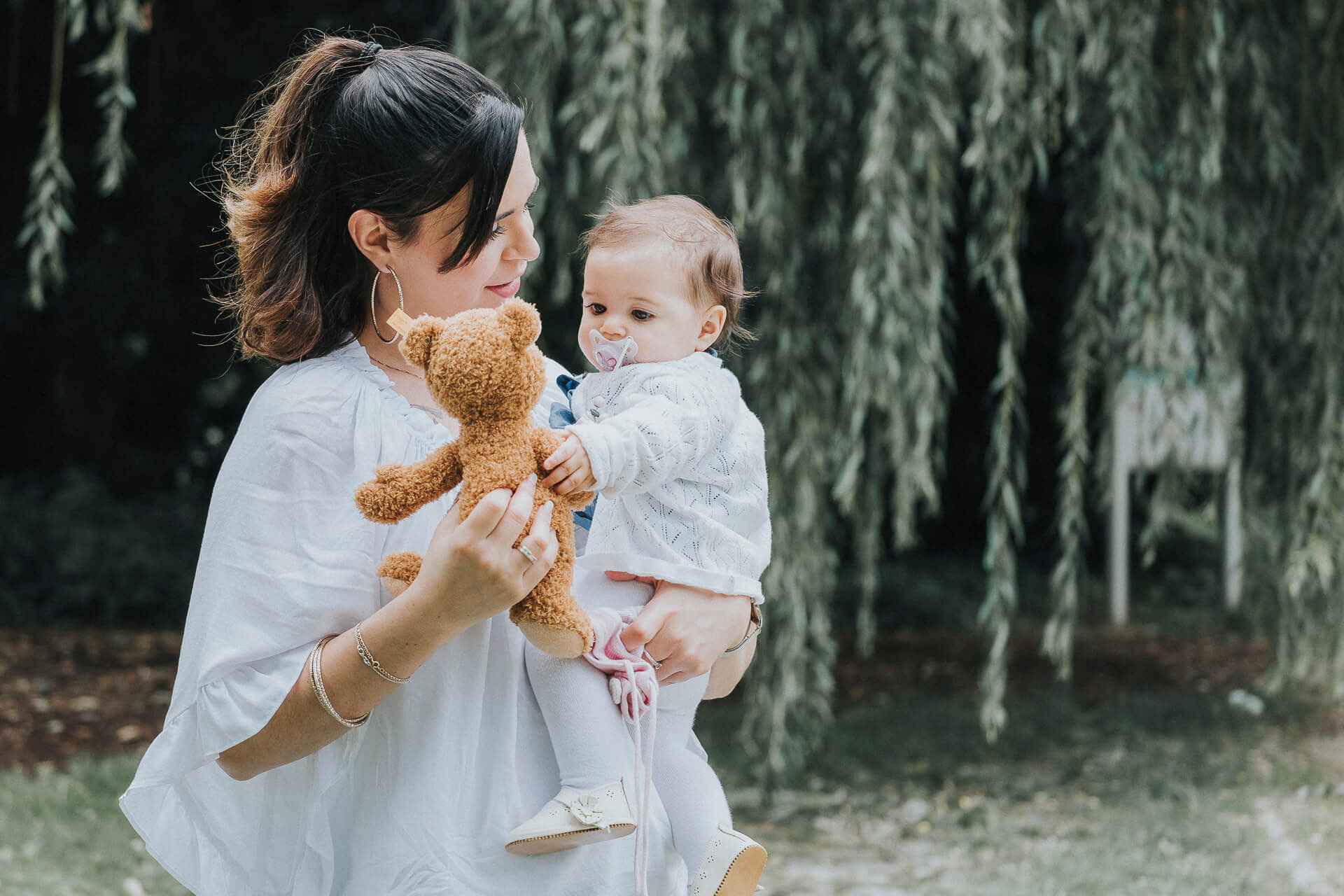 Mutter hält ihr Baby mit Stofftier im Arm vor einem grünen Baum.