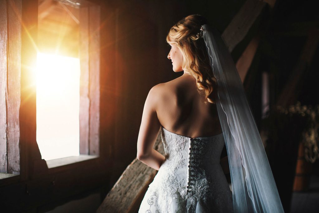 Braut im Hochzeitskleid steht im warmen Fensterlicht.