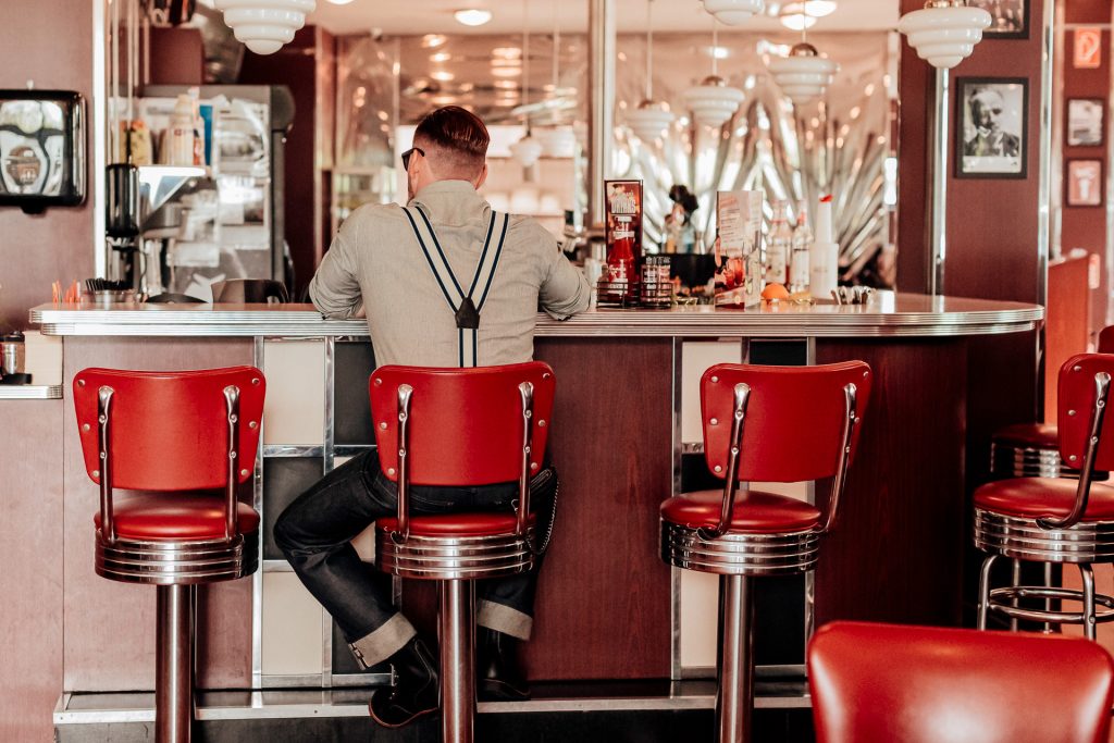 Bräutigam sitzt von hinten an der Theke eines American Diners mit roten Barhockern.