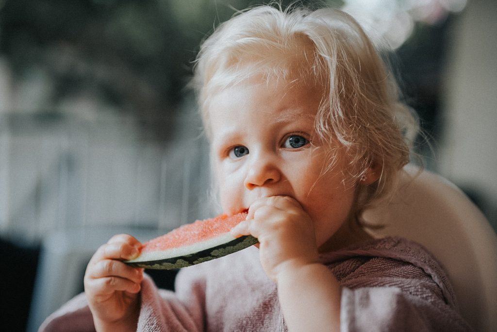 Blondes Kleinkind isst bei einem natürlichen Sommerporträt eine Scheibe Wassermelone.