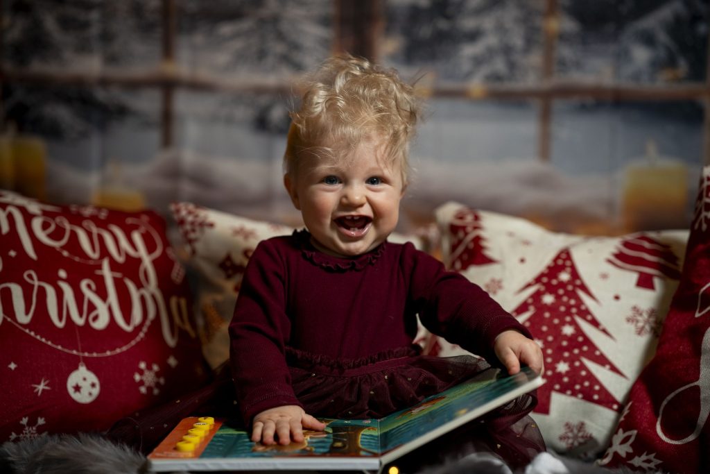 Lachendes Kleinkind sitzt mit einem Bilderbuch zwischen weihnachtlichen Kissen.