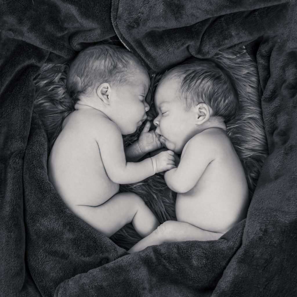 Zwei neugeborene Babys liegen einander zugewandt auf einer weichen dunklen Decke.